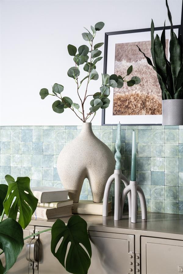 Rustieke vaas met groene eucalyptus takken en decoratieve kaarsen op een wit dressoir, wand met groene tegels, moderne planten en kunstwerk voor een natuurlijke en stijlvolle interieur look.