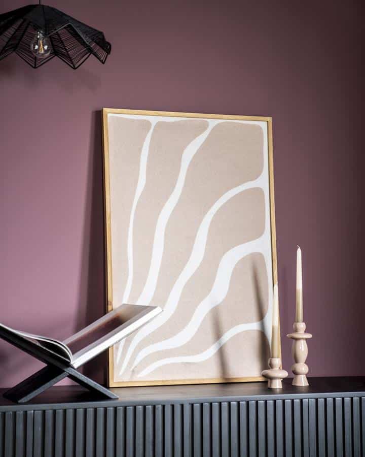 Eettafel met zwart frame en open boek, decoratief canvas met beige en witte abstracte organische lijnen en twee stijlvolle witte kandelaars op een donkergrijze meubel tegen een paarse muur.