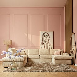 Lichte vintage bank in beige corduroy stof met strak design, comfortabele zithoek en decoratieve elementen zoals een grote spiegel en kunstzinnig portrait op een roze muur.