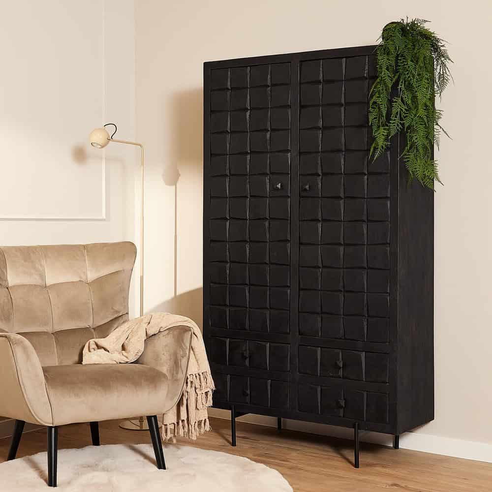 Houten zwarte kast met 8 lades in een modern interieur, gecombineerd met een beige velvet fauteuil, beige plaid, wandlamp en groen plantenaccent voor een stijlvolle en sfeervolle woonkamer.