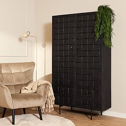 Houten zwarte kast met 8 lades in een modern interieur, gecombineerd met een beige velvet fauteuil, beige plaid, wandlamp en groen plantenaccent voor een stijlvolle en sfeervolle woonkamer.