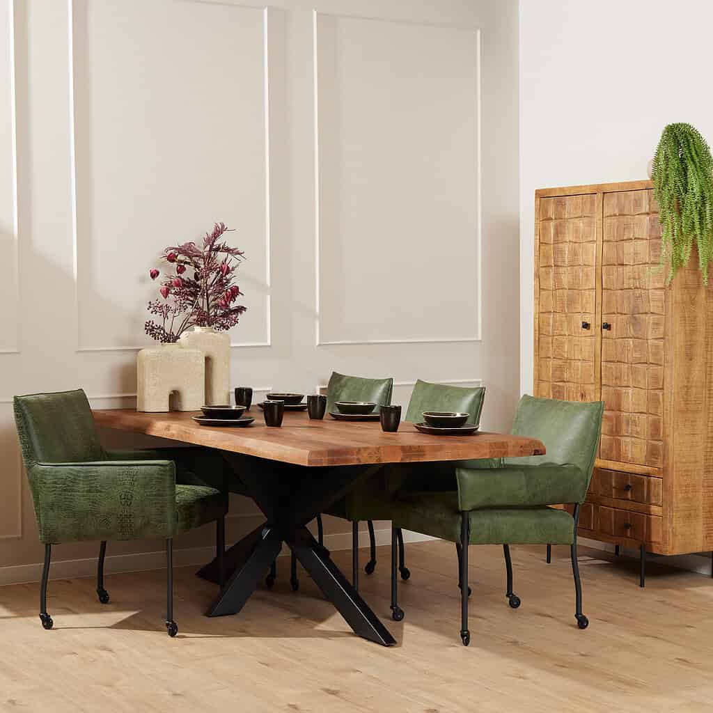 Eetkamertafel met zwarte X-poot en groene kuipstoelen in een modern interieur met houten kasten en decoratieve planten.