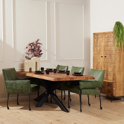 Eetkamertafel met zwarte X-poot en groene kuipstoelen in een modern interieur met houten kasten en decoratieve planten.