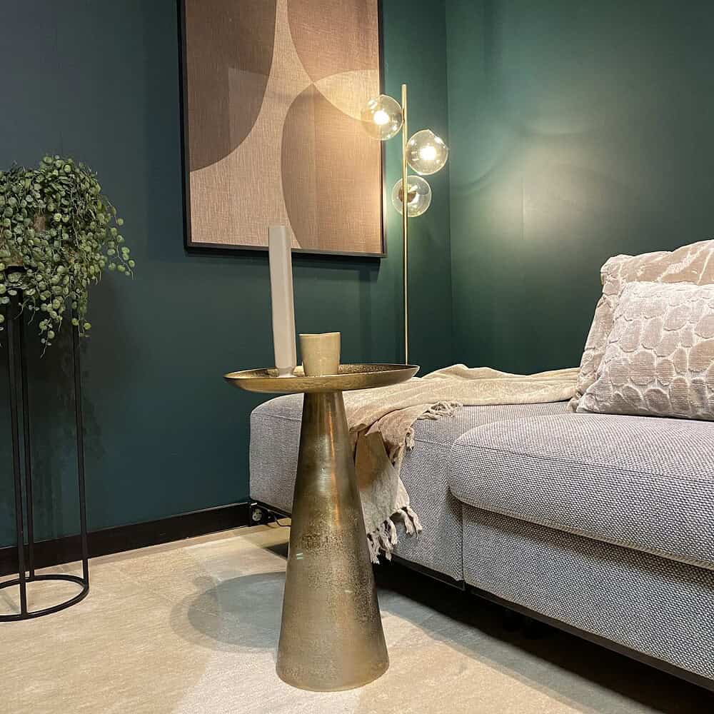Met goudkleurige bijzettafel, keramieken vaas en sfeerlichte met groene muur en modern kunstwerk, straalt de woonkamer elegantie en stijl uit. Perfect voor een warm en stijlvol interieur met Mokana meubelen.