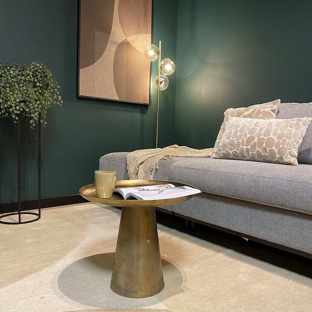 Gouden salontafel met decoratief accessoire en kopje, in modern interieur met groene wand, bank met textuurkussens, kunstwerk, en vloerlamp voor elegante woonstijl.