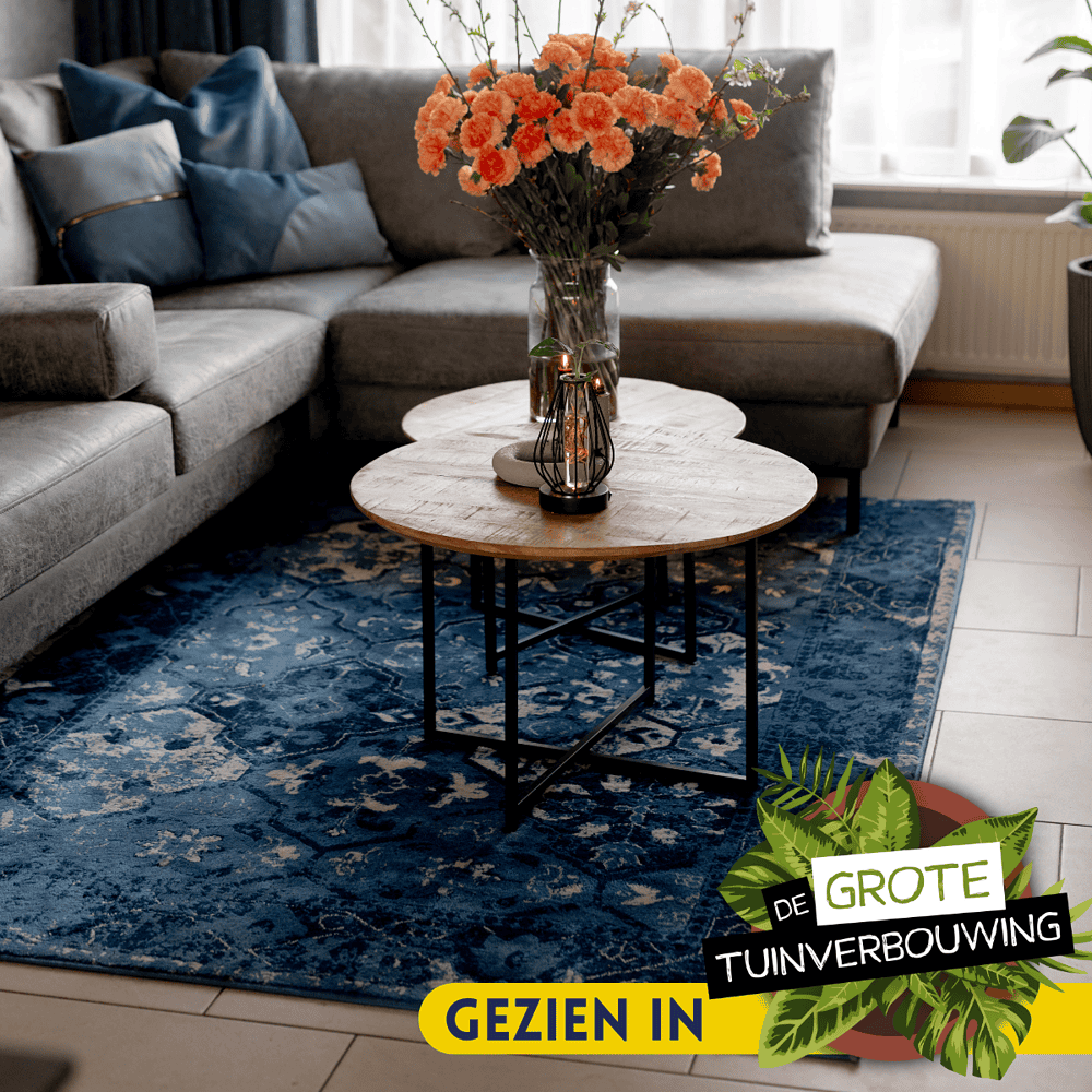 Naast de bank staan twee ronde salontafels met houten tops en zwart metalen onderstellen, decoratief ingericht met bloemen, kaarsen en sierstenen.