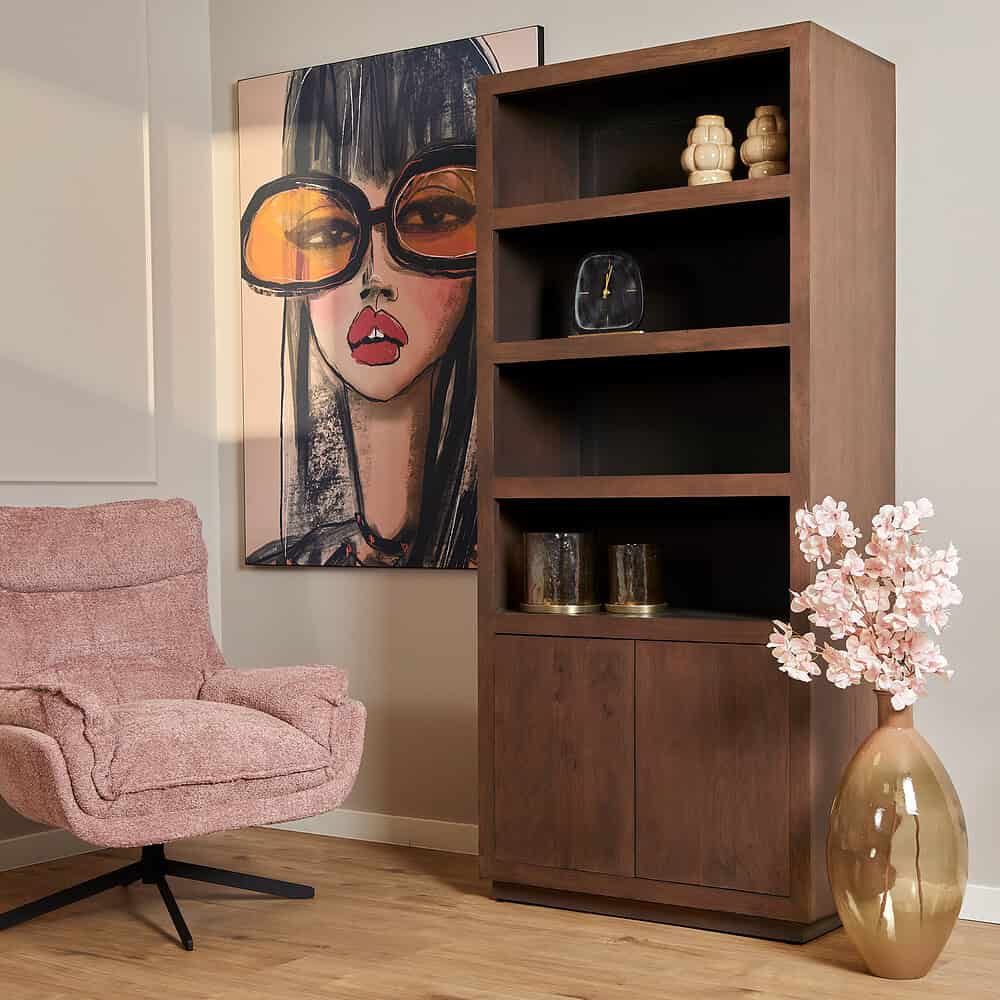 Vintage houten boekenkast met opbergvakken en decoratieve accessoires, gecombineerd met een chic roze fauteuil en een kunstzinnig portret voor een stijlvolle woonkamer.