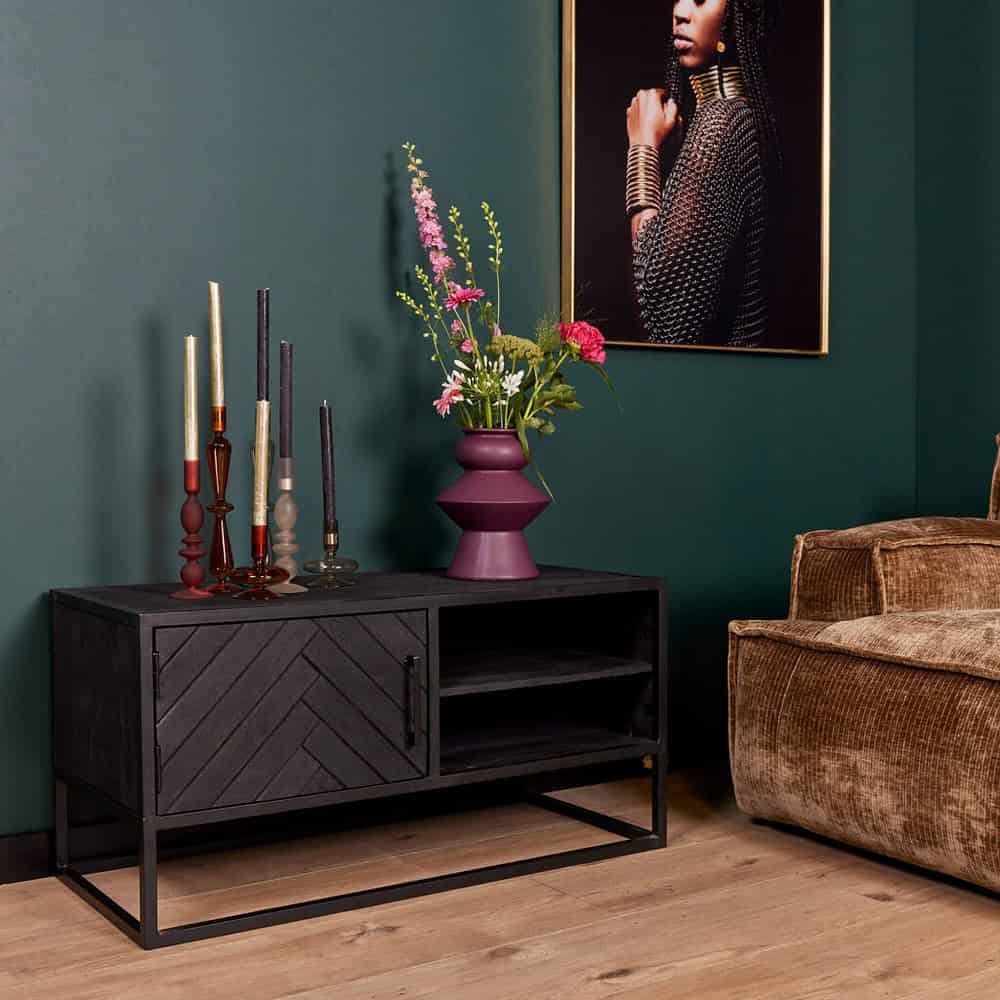 Kleine zwarte houten tv-meubel met metalen frame en open opberglade, met kleurrijke sieraden en bloemen op de bovenkant, in een modern interieur met groene muur en kunstwerk.