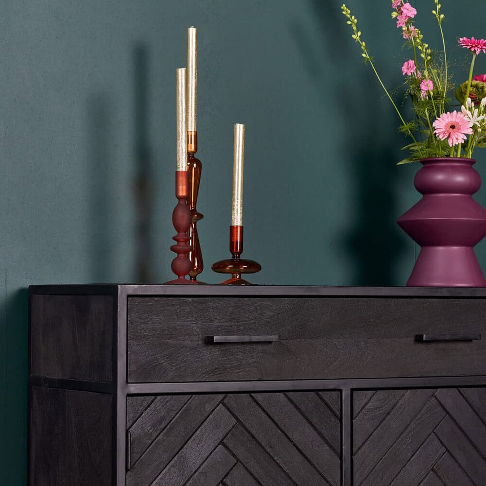 Kandelaar van gekleurd glas op zwart dressoir met bloemenvaas en roze bloemen, modern interieur accessoires, stijlvolle woondecoratie, elegant design meubels, Mokana Meubelen Nederland.