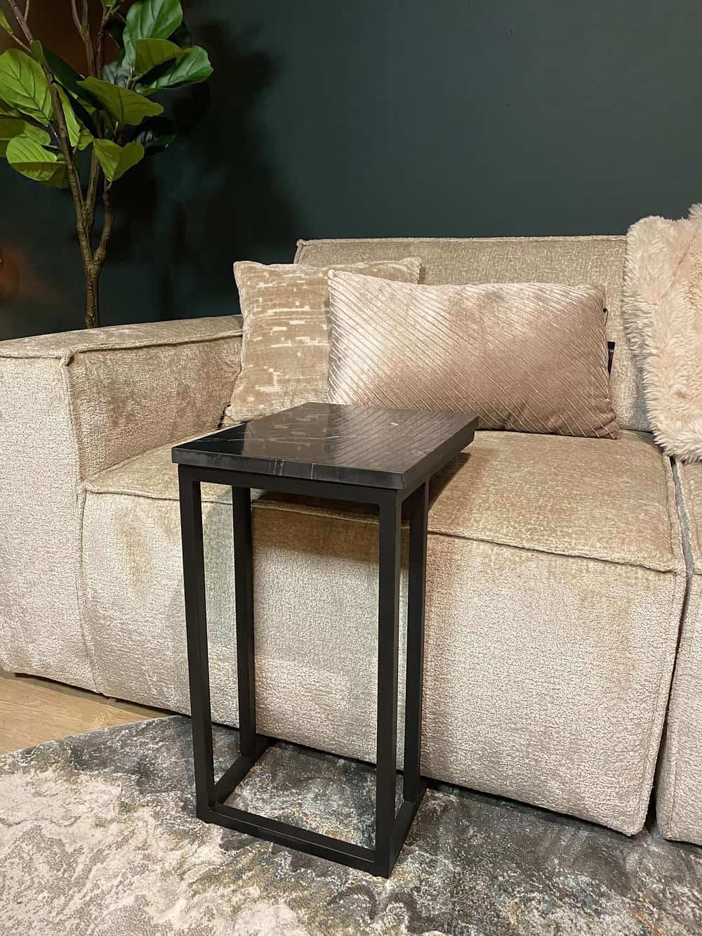 Stijlvolle zwarte bijzettafel met marmeren blad naast een beige stoffen bank, elegant interieur met zachte kussens en groene kamerplant op donkere muurachtergrond.