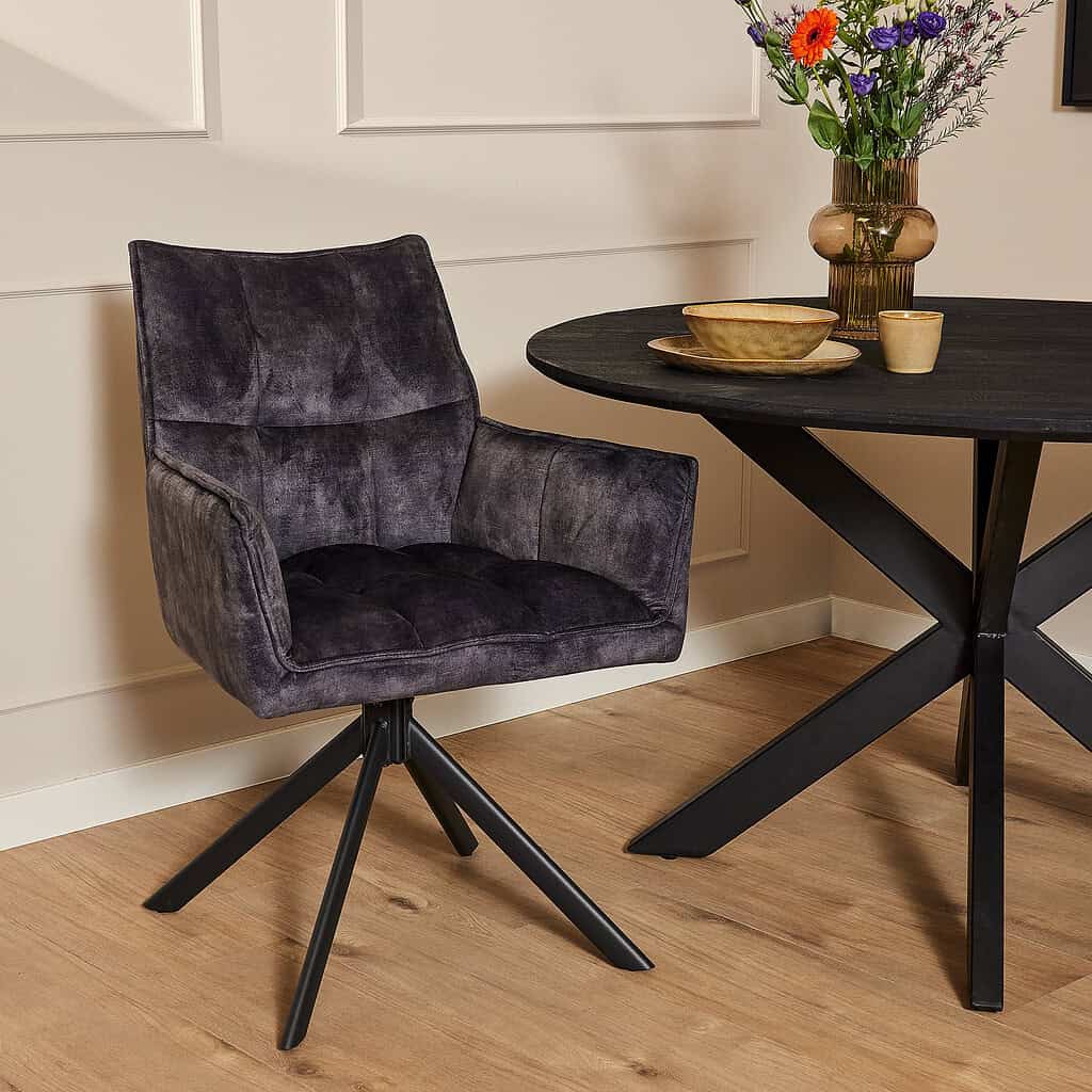 Metaal ogendoek fauteuil met gestoffeerde rugleuning en armleuningen, modern design met zwart metalen poot in een interieur met houten vloer en decoratieve tafel en bloemen.