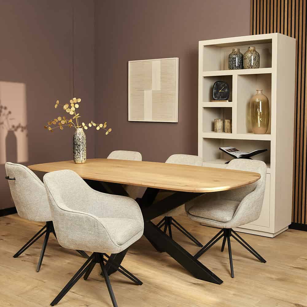 Modern eettafel met licht houten blad en zwarte X-vormige poot, omringd door comfortabele beige stoelen, met decoratieve vazen en een zachte warme sfeer.