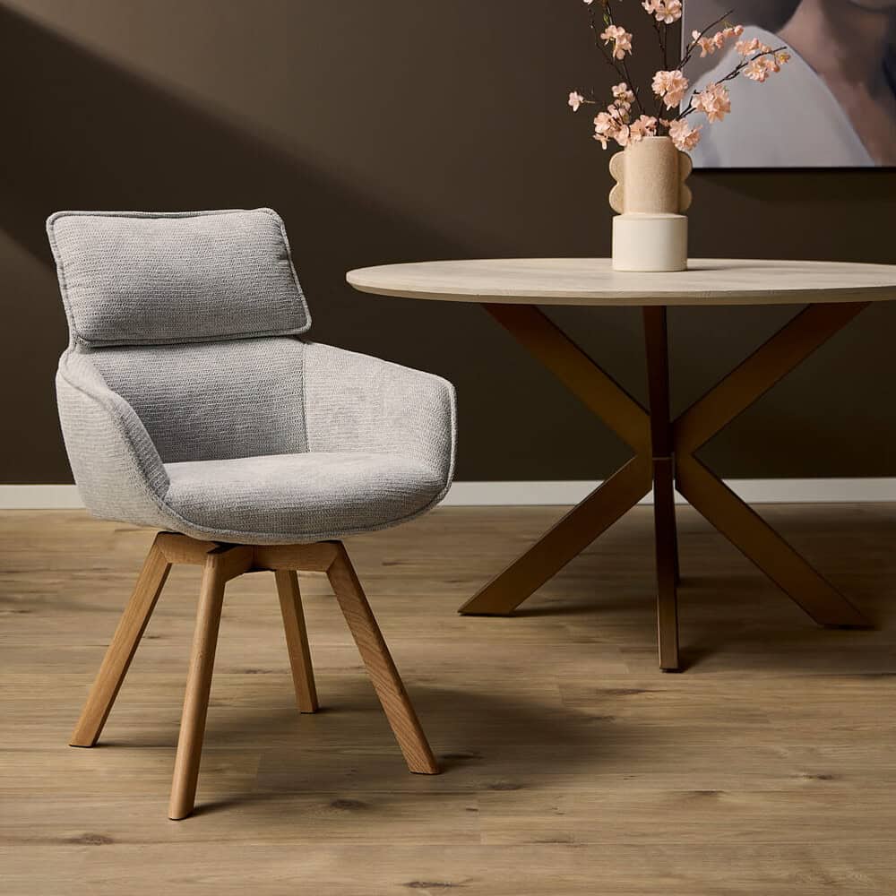 Modern fauteuil met lichtgrijze bekleding en houten poten, gecombineerd met een ronde eettafel met een minimalistisch design en een decoratieve vaas met roze bloemen, passend bij een eigentijdse woonstijl.