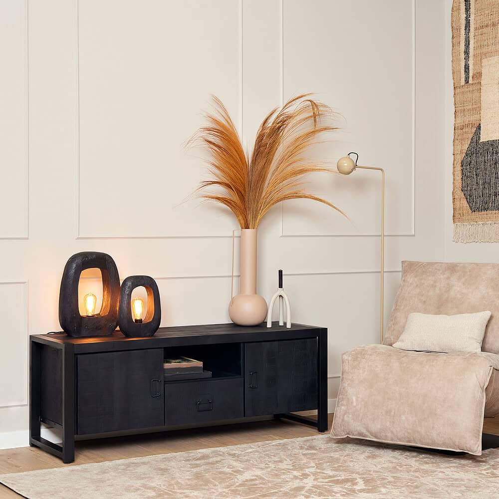 Zwarte houten tv-meubel met twee lades en open vakken, decoratief met sfeervolle lampen en grote vaas met beige decoratie, in een stijlvol, modern interieur.