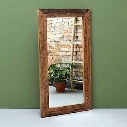 Houten spiegel van Mokana meubelen met natuurlijke afwerking, geplaatst tegen een groene muur, met een zicht op een binnenplaats met een plantenpot en een ladder.