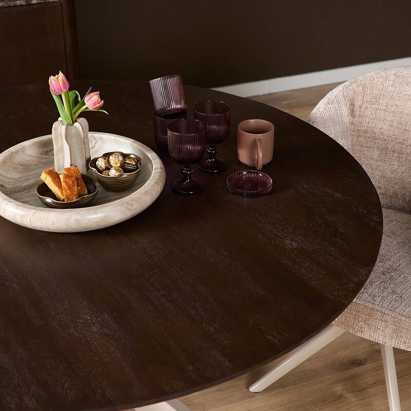 Eettafel Madison Brown met ronde vorm, 130 cm, met zandkleurige poot, afgebeeld met decoratieve glazen en bloemen.