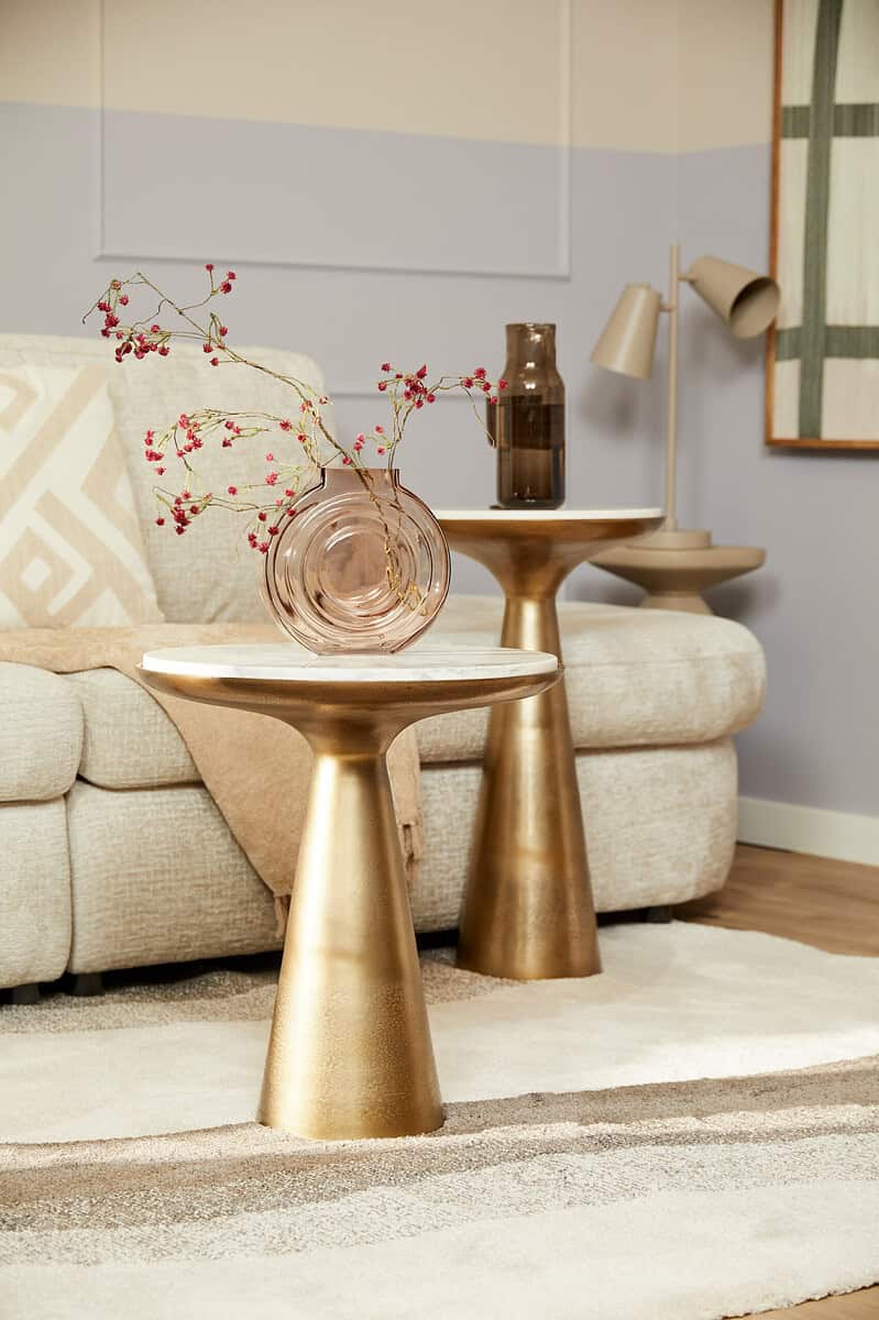 Moderne woonkamer met twee gouden bijzettafels, een vaas met takken en decoratieve accessoires, lichte muur en raam voor natuurlijke lichtinval.