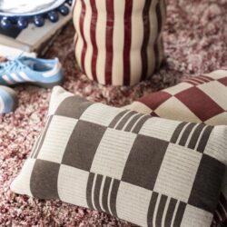 Decoratief kussen met geometrisch patroon en een keramische vaas met verticale strepen, geplaatst op een zachte, roze tapijtvloer voor een stijlvolle interieurdecoratie.