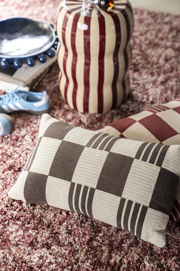 Decoratief kussen met geometrisch patroon en een keramische vaas met verticale strepen, geplaatst op een zachte, roze tapijtvloer voor een stijlvolle interieurdecoratie.