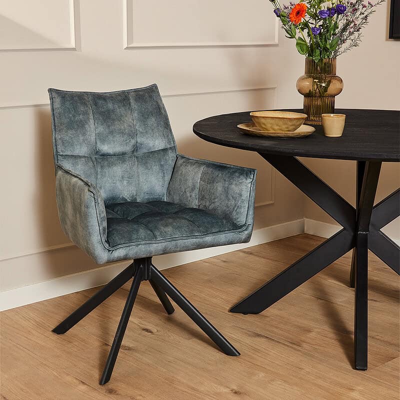 Duidelijke afbeelding van de Mokana Dani Velvet eetkamerstoel in blauw met armleuning, geplaatst naast een zwart eettafel met decoratieve vazen en bloemen.