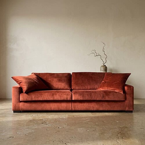 Luxe fluwelen bank in een warme roestkleur met bijpassende kussens, geplaatst in een minimalistische woonkamer met een neutrale muur en een decoratieve vaas op de achtergrond.