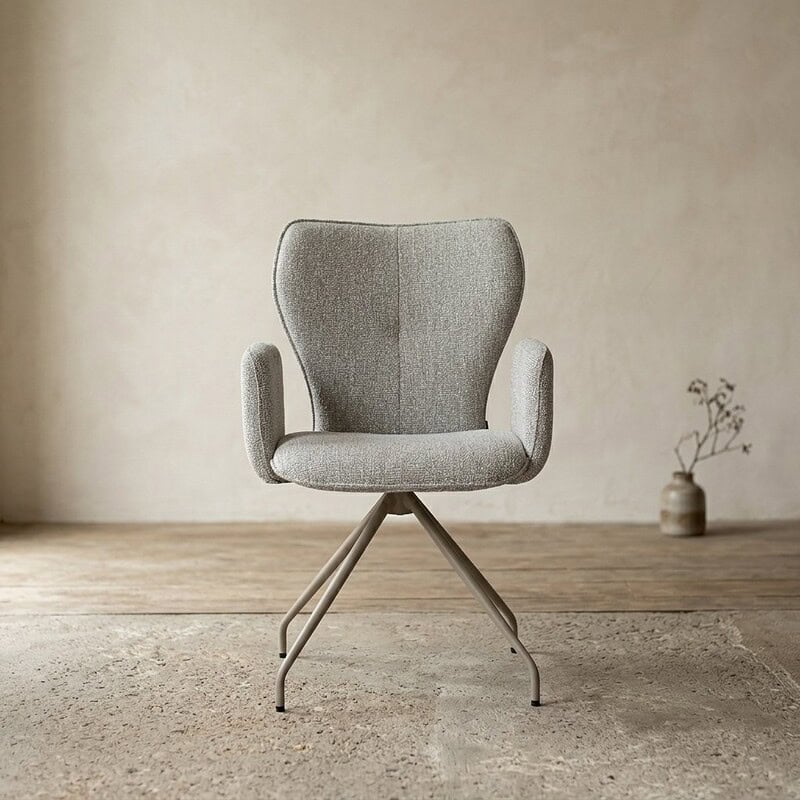 Moderne Mokana designstoel met comfortabele armleuningen en metalen onderstel, geplaatst in een minimalistisch interieur met een vaas en plant op de achtergrond.