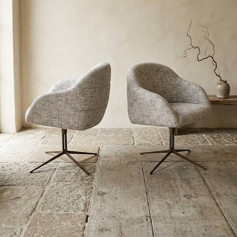 Twee moderne stoffen lounge stoelen met draaibare onderstellen, geplaatst op een landelijke stenen vloer in een minimalistische interieursetting.