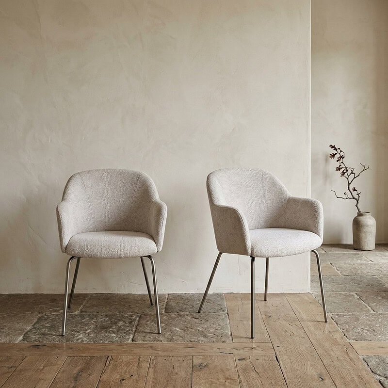 Twee lichte stoffen fauteuils met metalen poten in een minimalistische woonkamer, gecombineerd met een decoratieve vaas en takken voor een elegante uitstraling.