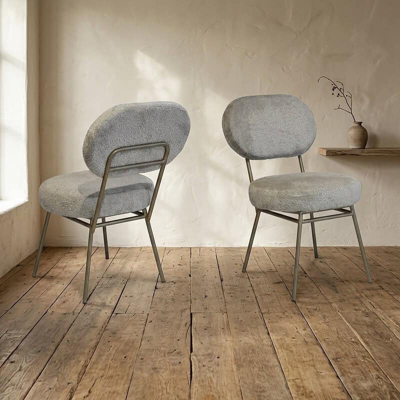 Twee moderne houten stoelen met zachte, lichtgrijze bekleding in een minimalistische kamer met houten vloer en een eenvoudige vaas op de achtergrond.