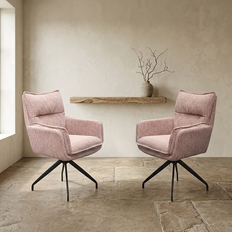 Twee moderne, zachte roze fauteuils met zwarte metalen poten in een minimalistische woonkamer met een houten plank en een vaas met takken op de achtergrond.