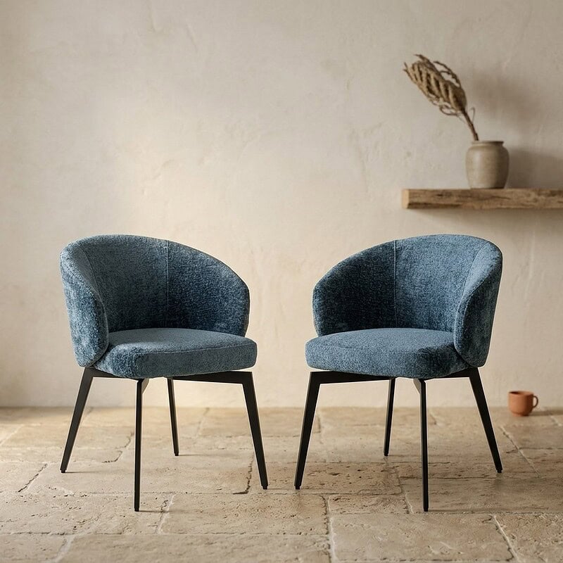 Zwei moderne blaue Sessel mit schwarzen Beinen in einem minimalistischen Wohnzimmer, im Hintergrund ein Holzregal und eine Vase mit Trockenblumen.