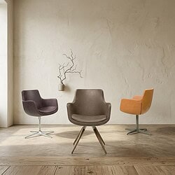 Drie stijlvolle moderne fauteuils in verschillende kleuren op een houten vloer, met een eenvoudige muur en decoratief takken in een vaas op de achtergrond.