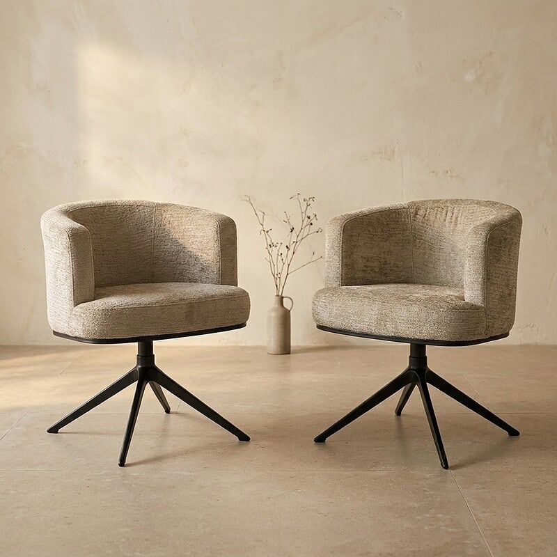 Twee moderne fauteuils met zachte bekleding en zwarte poten, geplaatst in een minimalistische interieursetting met een vaas met takken op de achtergrond.