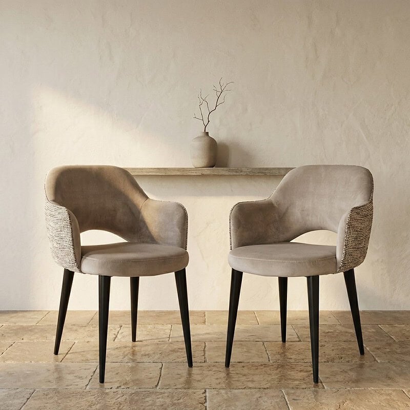 Zwei moderne beigefarbene Esszimmerstühle mit weicher Polsterung und Holzbeinen, platziert in einem minimalistischen Interieur mit einer hellen Wand und einer dekorativen Vase im Hintergrund.