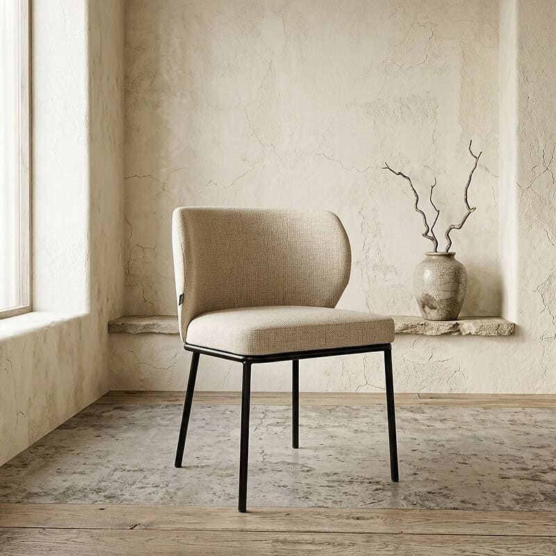 Moderne eetkamerstoel met beige stoffering en zwarte metalen poten, geplaatst in een minimalistische interieursetting met een vintage vaas en takken op de achtergrond.