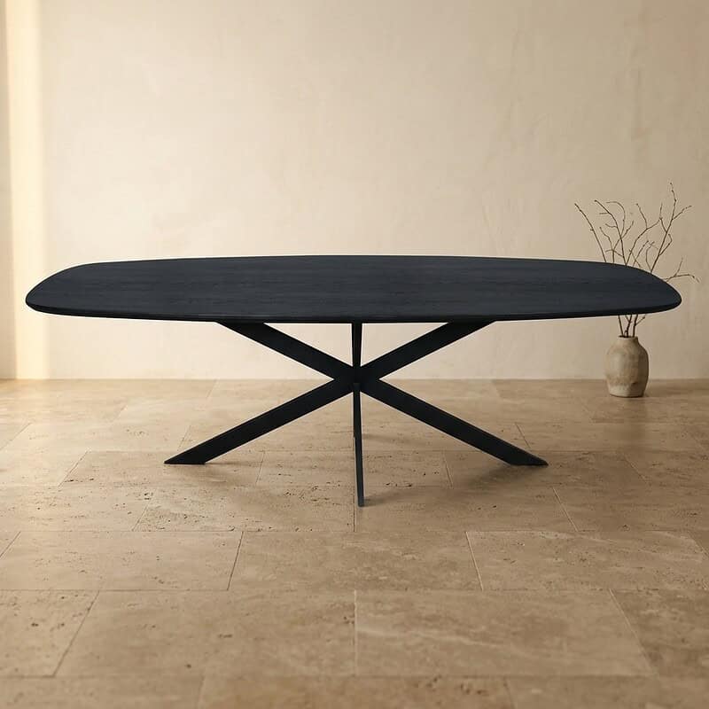 Moderne zwarte eettafel met uniek ontwerp in een minimalistische interieurstijl, geplaatst op een lichte houten vloer, met een decoratieve vaas en takken op de achtergrond.