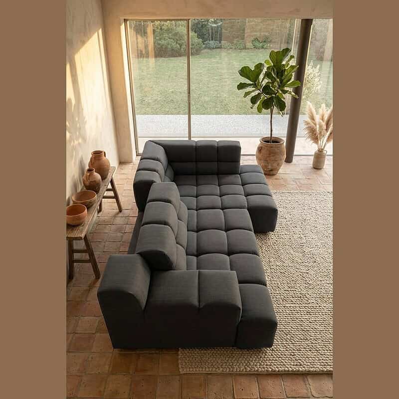 Moderne L-vormige bank in donkergrijze stof, perfect voor een stijlvolle en comfortabele zithoek in een woonkamer met grote ramen en natuurlijke decoratie.