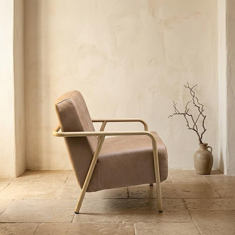 Moderne fauteuil met beige bekleding en metalen armleuningen, geplaatst in een minimalistische interieursetting met een keramieken vaas en een tak in de achtergrond.