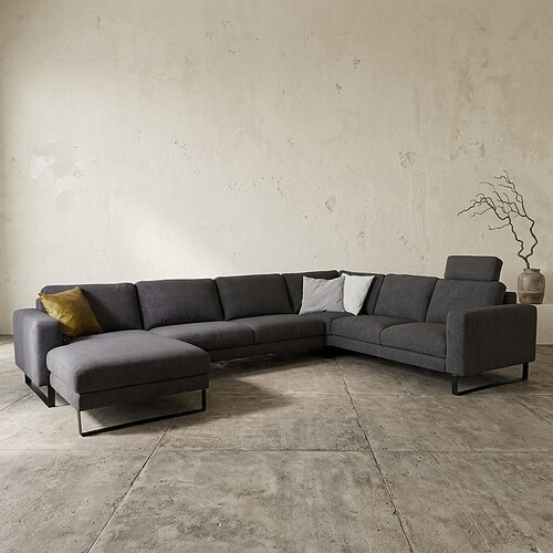 Moderne Loungebank in een minimalistische woonkamer met een grijze stofafwerking, gecombineerd met sierkussens en een decoratieve plant op de achtergrond.