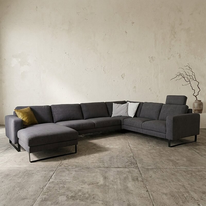 Ein modernes Loungesofa in einem minimalistischen Wohnzimmer mit grauem Stoffbezug, kombiniert mit Zierkissen und einer dekorativen Pflanze im Hintergrund.