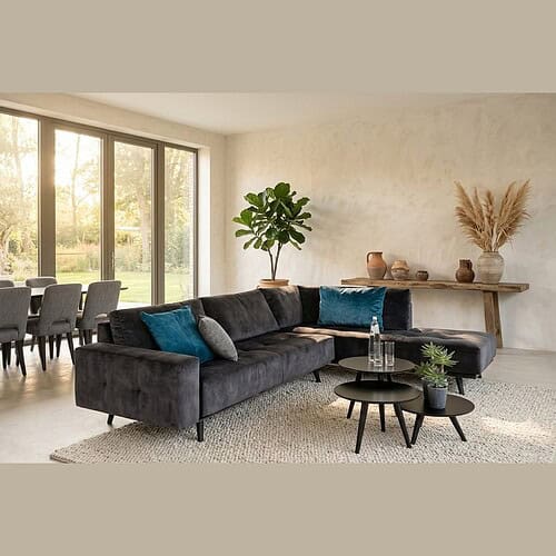 Modern zwart stoffen hoekbank met blauwe en grijze kussens in een lichte woonkamer met grote ramen en natuurlijke decoratie. Zeer stijlvolle en comfortabele hoekbank, perfect voor een moderne woonkamer.