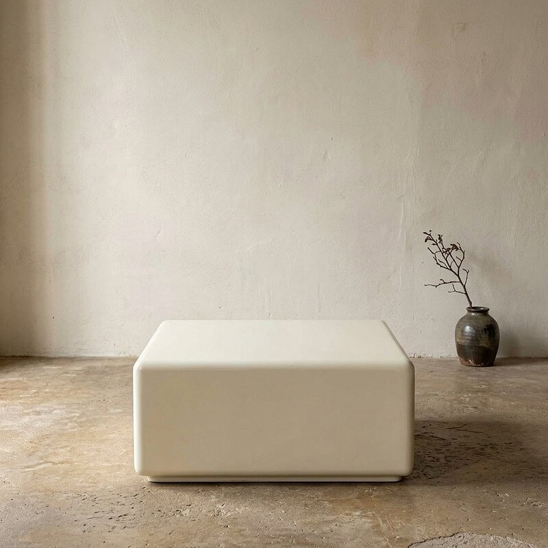 Moderne witte bank in een minimalistisch interieur met een ruwe betonvloer en een eenvoudige vaas met takken op de achtergrond, perfect voor een strak en eigentijds woonconcept.