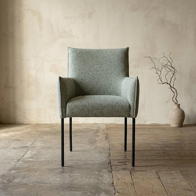 Moderne houten armstoel met zachte bekleding in een minimalistisch interieur, geplaatst op een houten vloer met een decoratieve vaas en takken op de achtergrond.