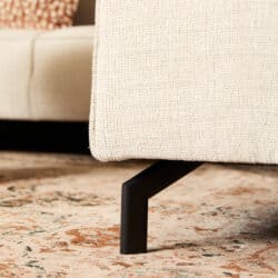 Close-up van een beige stoffen bank met zwarte metalen poot, geplaatst op een traditioneel tapijt in een woonkamer.