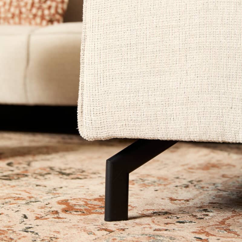 Close-up van een beige stoffen bank met zwarte metalen poot, geplaatst op een traditioneel tapijt in een woonkamer.
